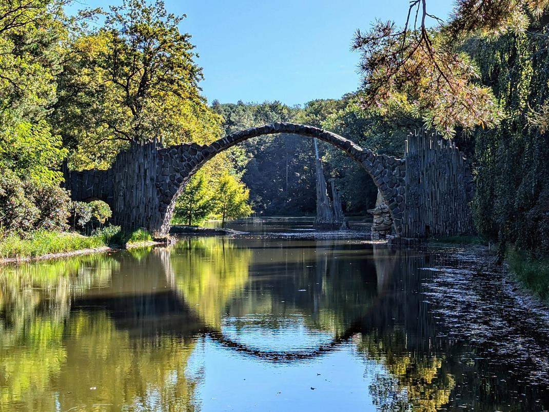 Rakotzbrücke in Gablenz, Kromlau umgeben von dichtem Herbstwald und dem ruhigen Rakotzsee, der das Bild der halbkreisförmigen Steinbrücke spiegelt.